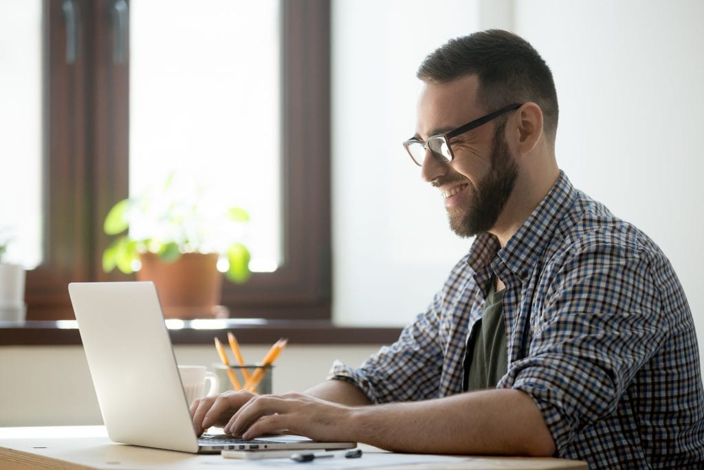 Happy male office worker smiling typing message at laptop